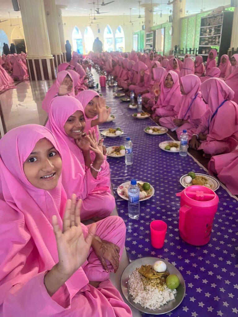 Offrir un repas :  Jeunes orphelines au Bangladesh partageant un repas chaud offert par l’association Les P’tits Villages d’Afrique, souriantes et vêtues d’un uniforme rose.