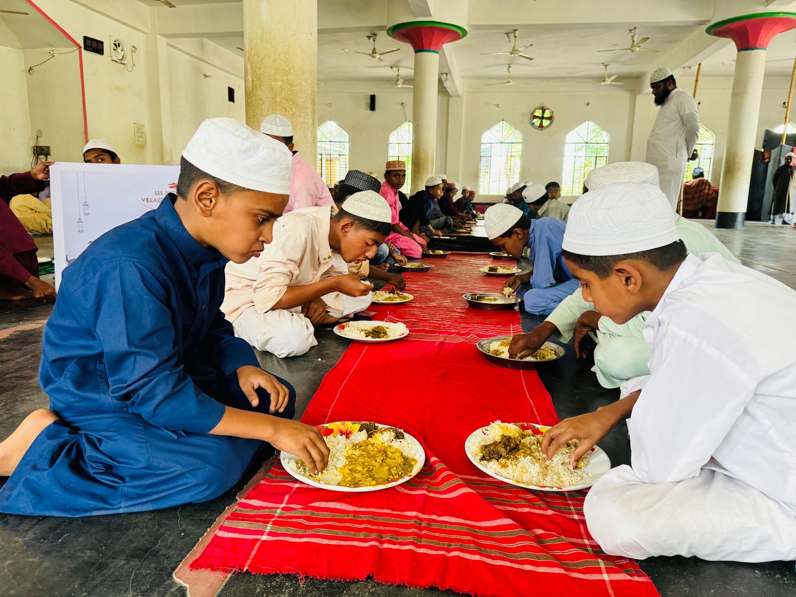 Enfants partageant un repas chaud distribué par l’association Les P’tits Villages d’Afrique, assis sur des tapis dans une salle communautaire.